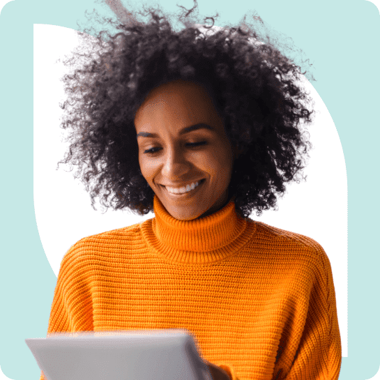 A woman with curly hair, wearing an orange sweater, smiling while looking at a tablet.